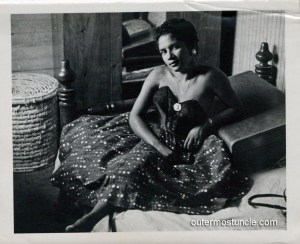 1950's black and white photo of a mysterious woman with an alluring look. She's black and wearing a cocktail dress.