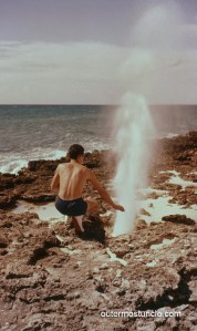 A 1950's photograph of a man, crouching next to a tidal hole in the coral cliff. Water is spewing out of it. San Salvador Island, Bahamas.