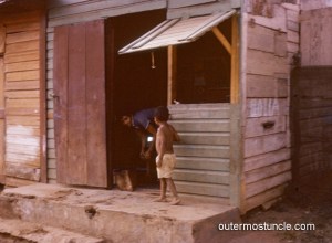 1950's Bahamas. A boy on a prch with a woman looking out the doorway.