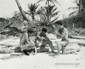 A 1950s black and white photograph of 3 men pretending tobe shipwrecked. They are trying to drink water from coconuts.