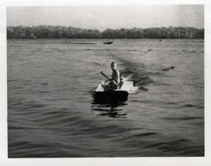 A black and white photo of a boy in a homemade, wooden boat.