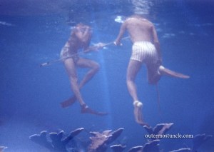 An underwater photo, looking up from below, of two young men at the surface. Just legs and shorts and a speargun.