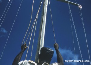 A Bahamian as viewed against the sky, from behind, operating the ropes of a sail.