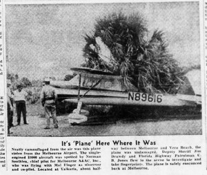 A black and white newspaper photo. An aircraft covered in palm fronds. 3 men stand in front of it.