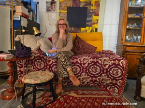 A woman sits, legs crossed, on a colorful sofa, smiling at the lens.