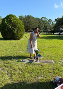 Angela, standing at her father's grave in a cemetery, covering her face with her hand.