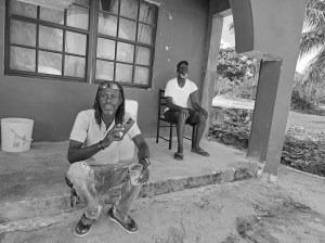 2 men on a porch in Mayaguana. One sits on a step and holds a plastic tumbler of gin and a cell phone. The other sits in a chair.